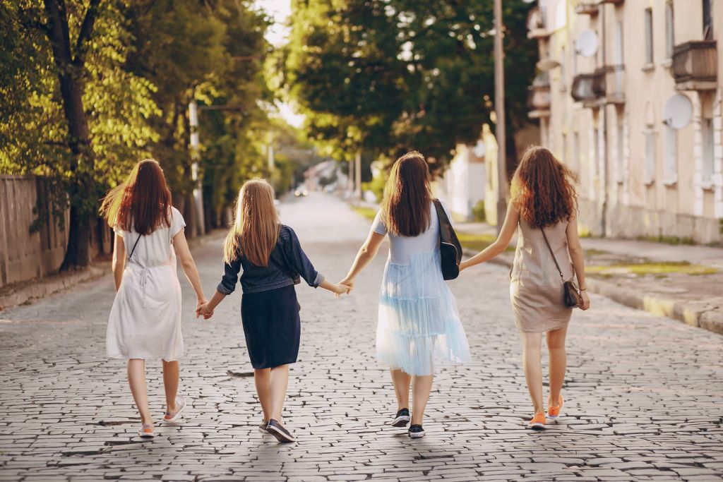 Four women walking hand in hand down a cobblestone street, surrounded by trees, in a sunny, relaxed setting.