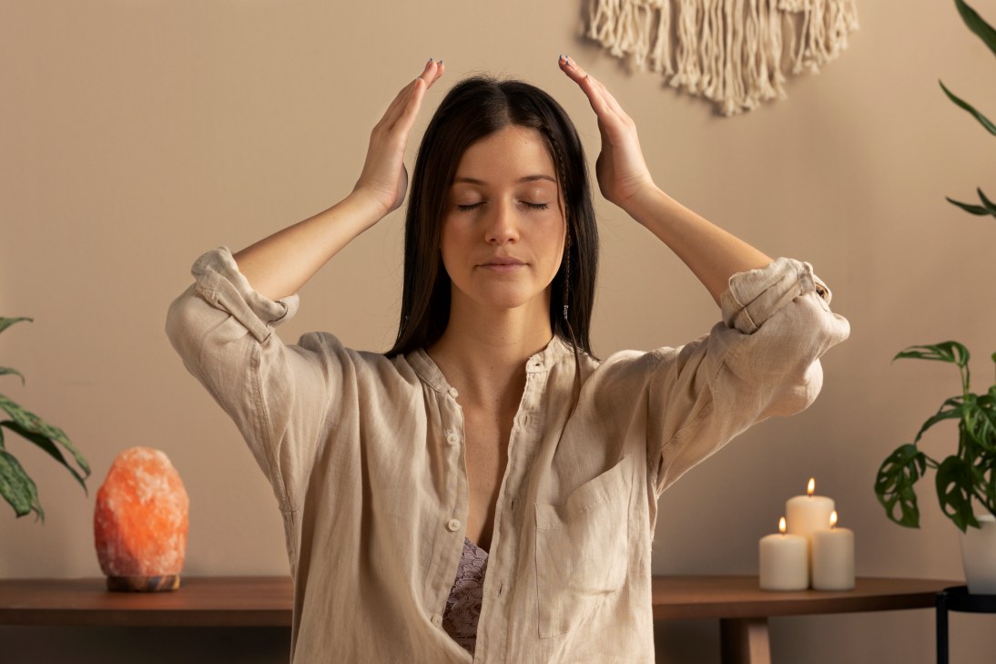 A woman with closed eyes meditates, hands near head, in a tranquil room with plants, candles, and a Himalayan salt lamp.