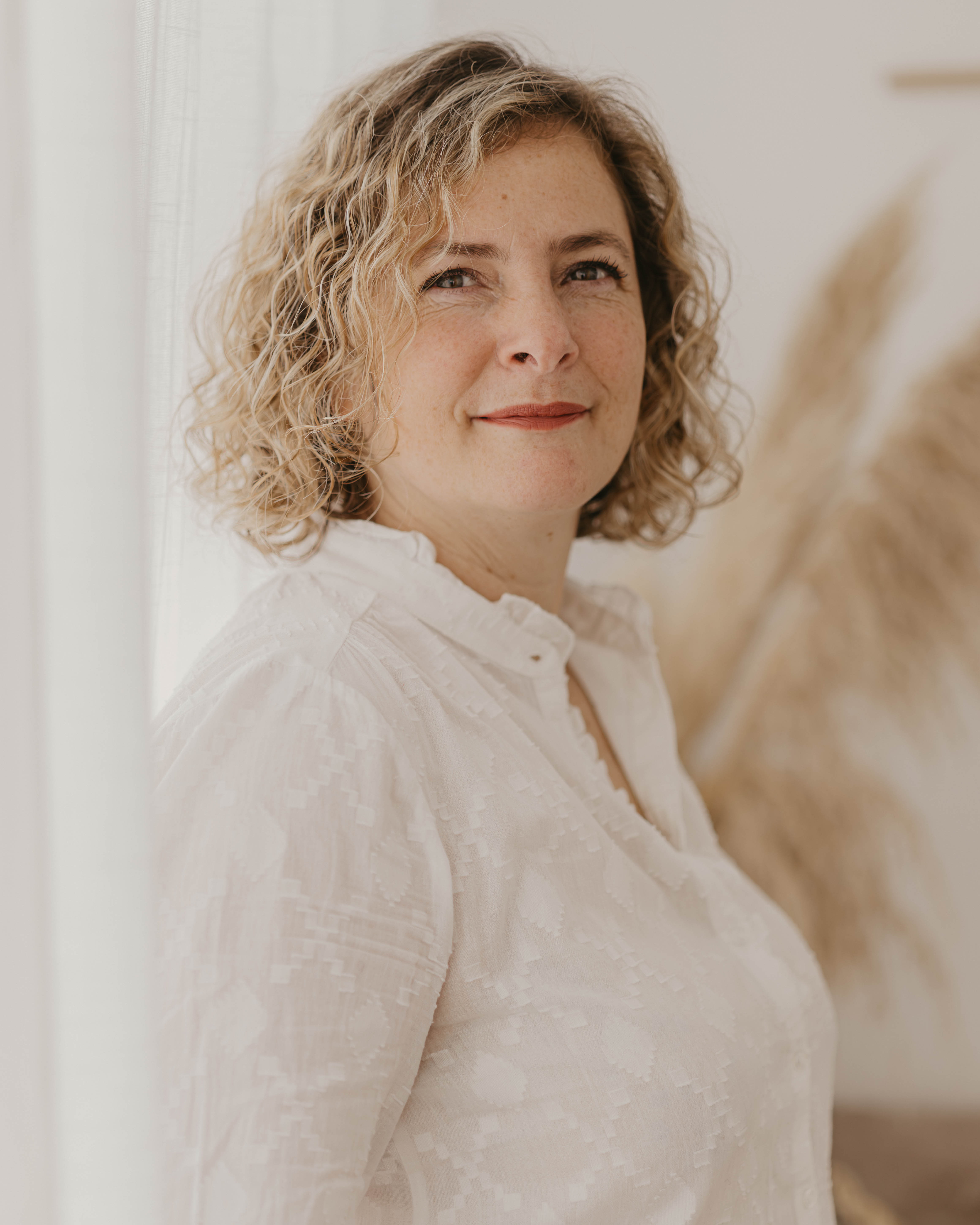 Woman with curly blonde hair in a white blouse, smiling softly, standing indoors near a window with soft lighting.