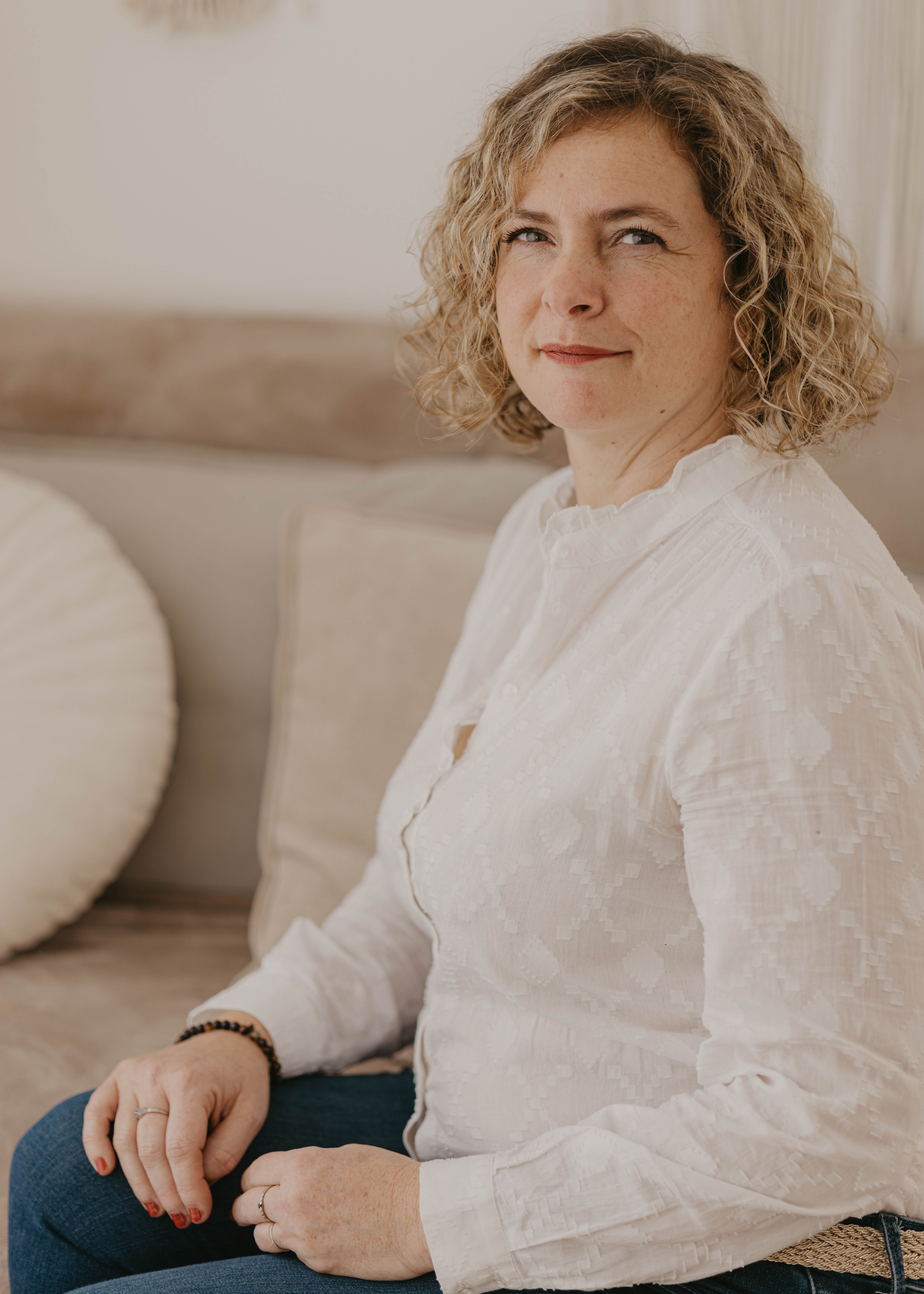 A woman with curly hair sits on a couch, wearing a white blouse and jeans, looking into the camera with a slight smile.